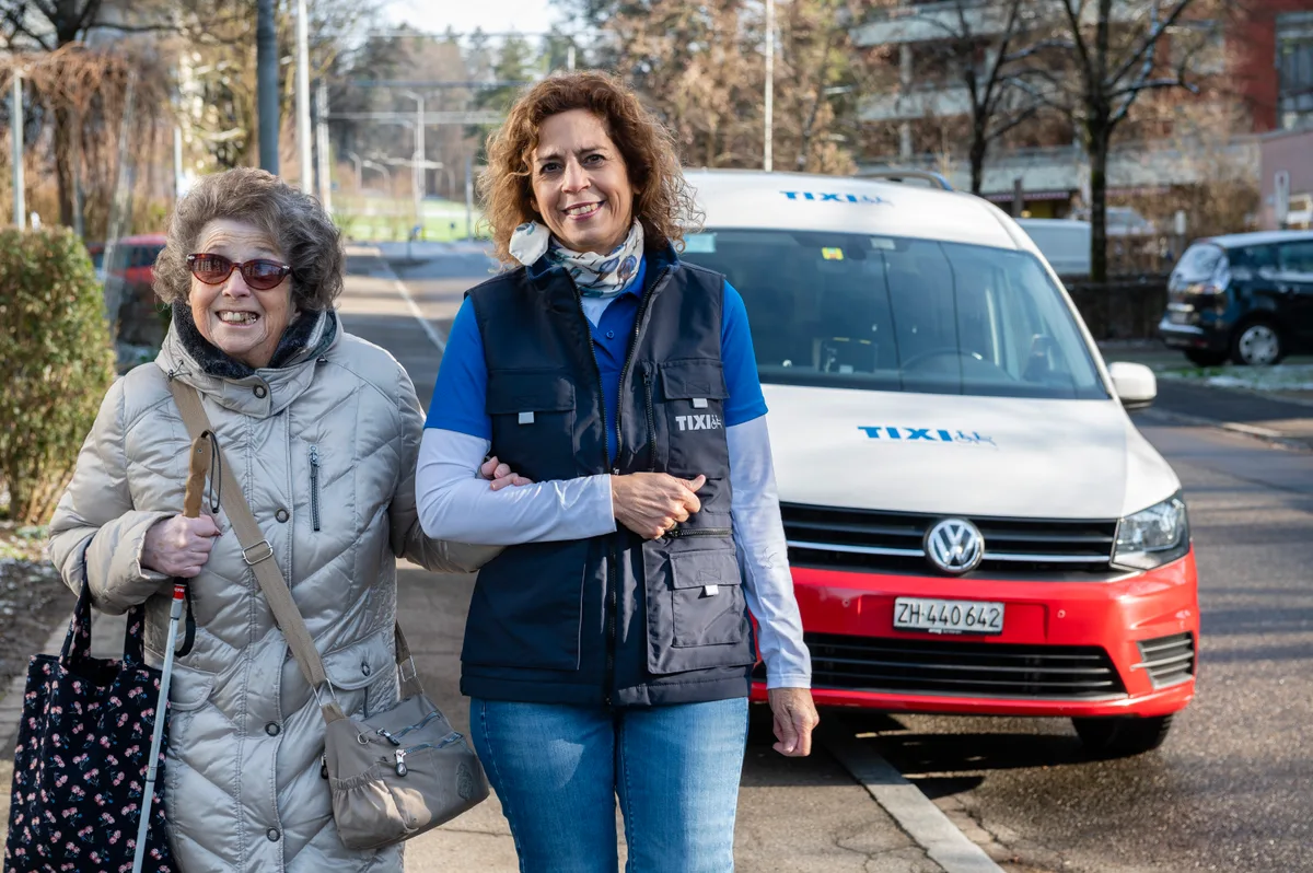 Eine Frau begleitet eine ältere sehbehinderte Frau vom Tixi-Auto weg.